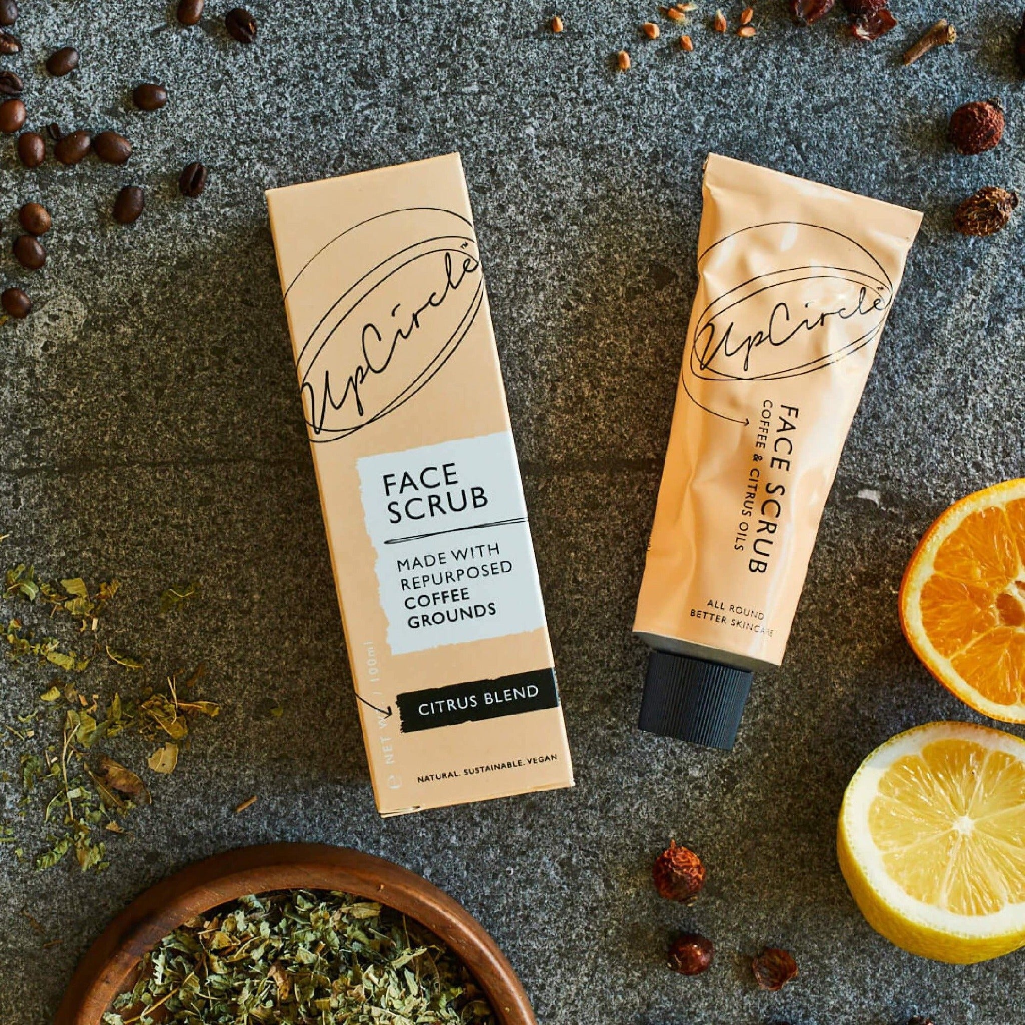 UpCircle face scrub of coffee and citrus oils aluminum tube with its package next to it and some spices and orange surrounding.